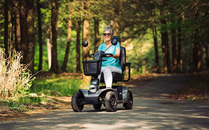 A woman with short silver hair is seated on a Pursuit 2 scooter on a path in the woods.